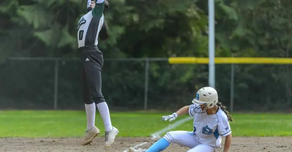 A softball player in a black uniform leaps to catch a ball as another player in blue slides into base, illustrating a close play that could invoke the mercy rule in baseball.
